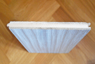 Das Bild zeigt den Queerschnit einer Dreischichtdiele. Ober -und Unterlage sind aus Edelholz . Das Mittelteil ist aus Nadelholz. Das Muster seht auf einem Parkettboden.